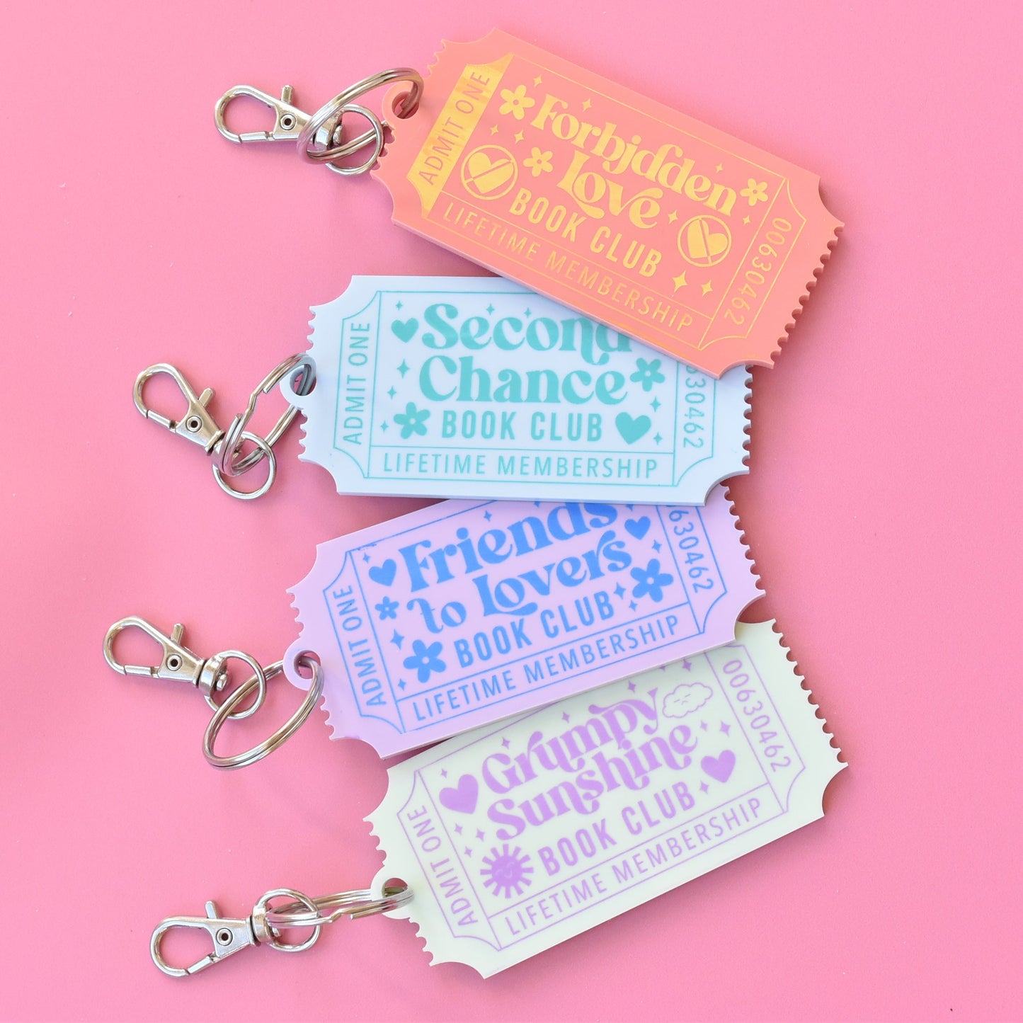 Set of four romance book trope keychains shaped like vintage tickets, featuring pastel colors and phrases like 'Forbidden Love,' 'Second Chance,' 'Friends to Lovers,' and 'Grumpy Sunshine' on a pink background
