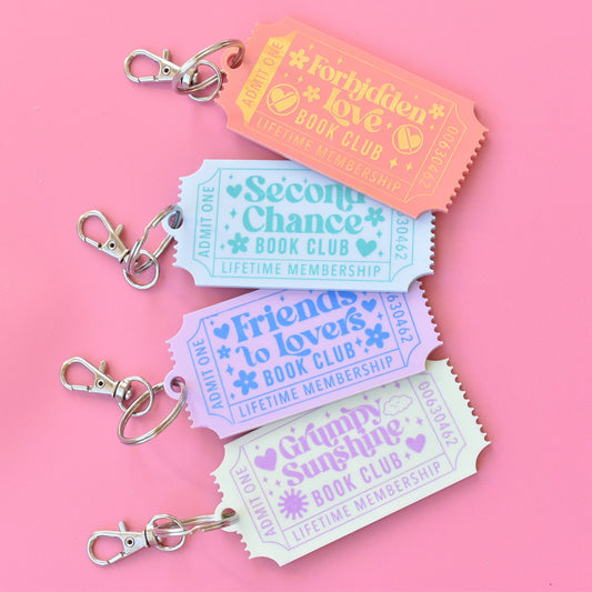 Set of four romance book trope keychains shaped like vintage tickets, featuring pastel colors and phrases like 'Forbidden Love,' 'Second Chance,' 'Friends to Lovers,' and 'Grumpy Sunshine' on a pink background